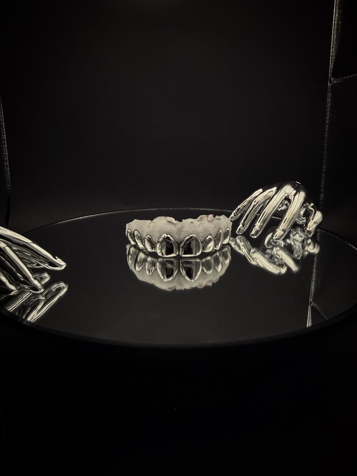 Verchromte Grillz von BRIGHTZ. zwischen zwei Chrom-Händen auf schwarzem Spiegel